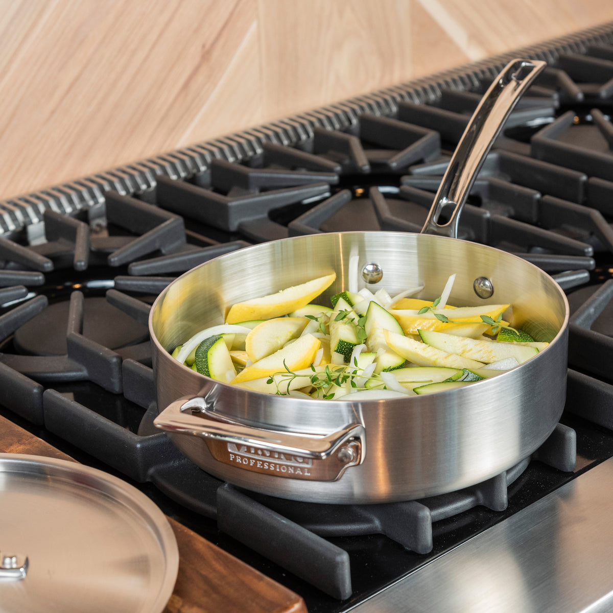 The Viking Professional 5-Ply Stainless Steel 3.4-Quart Sauté Pan by Viking Culinary Products, filled with sliced zucchini and onions, cooks on a gas stove while its metal lid rests on a wooden countertop.