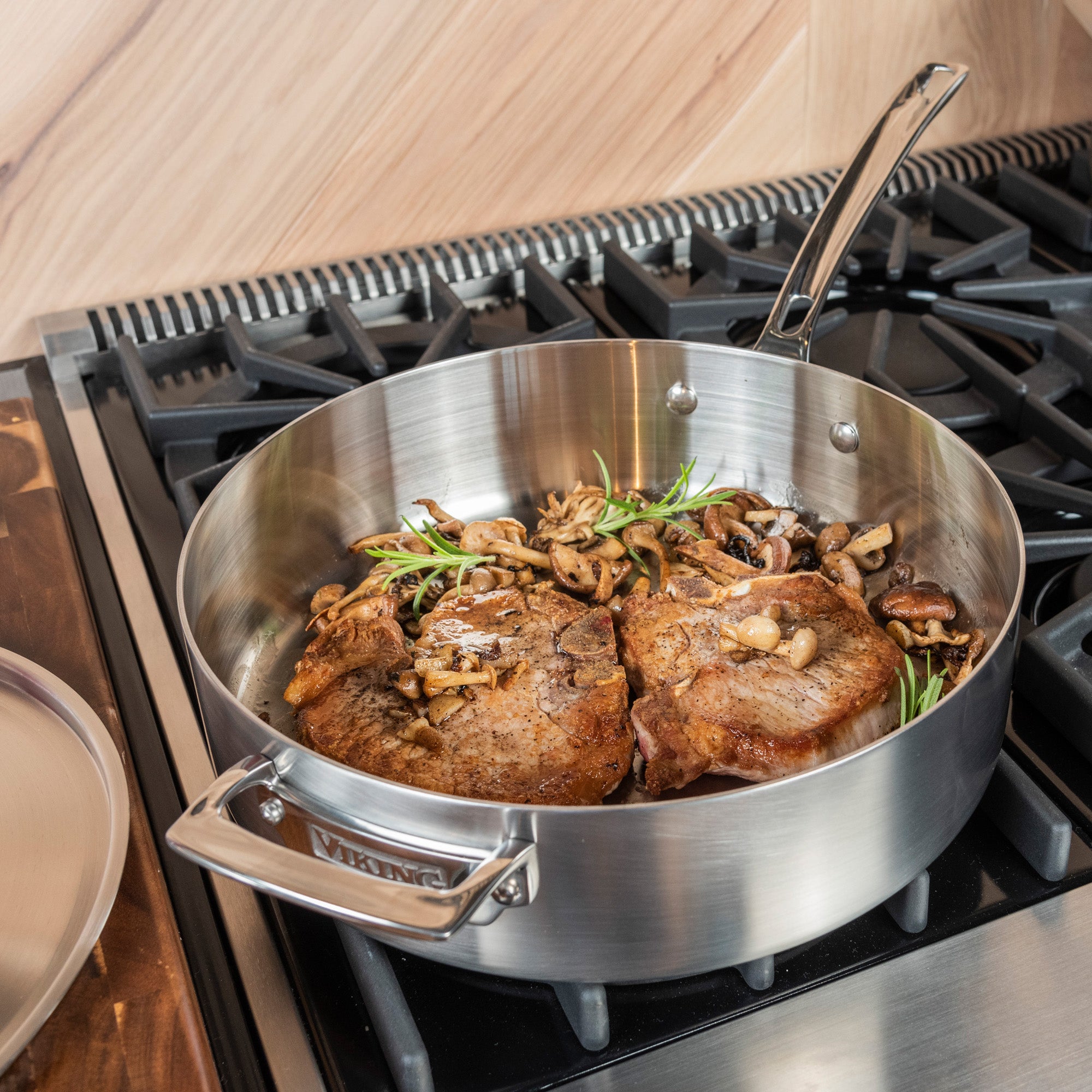 The Viking Professional 5-Ply Stainless Steel 6.4-Quart Sauté Pan with Metal Lid by Viking Culinary Products rests on a stovetop with sleek black grates, holding two cooked steaks, mushrooms, and rosemary. The made-in-USA pan is partially covered by its lid.