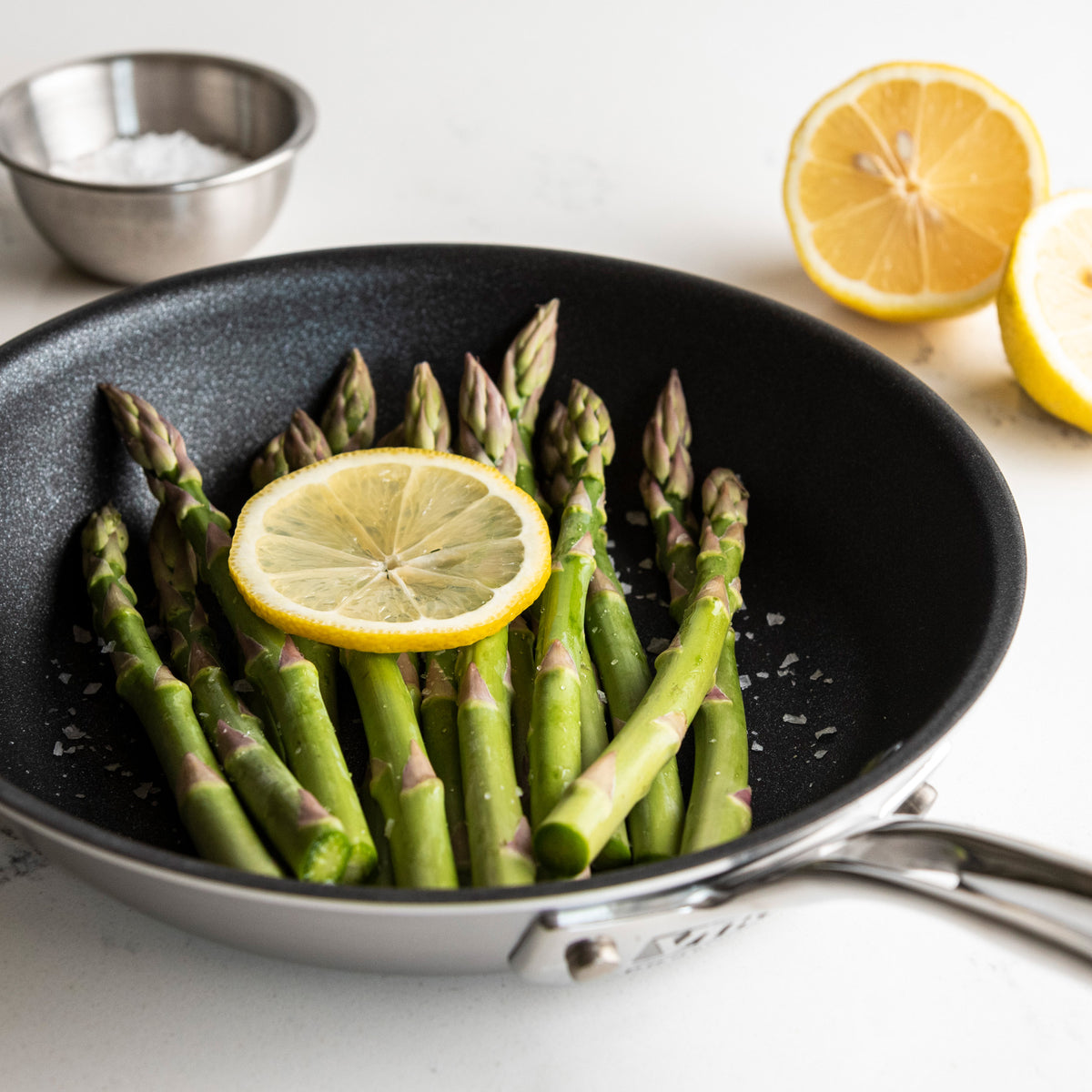 The Viking Culinary Products Professional 5-Ply 8-Inch Nonstick Fry Pan cooks fresh asparagus topped with lemon and salt. A halved lemon and bowl of salt sit nearby. Stay-cool handles offer comfort while cooking on a white surface.