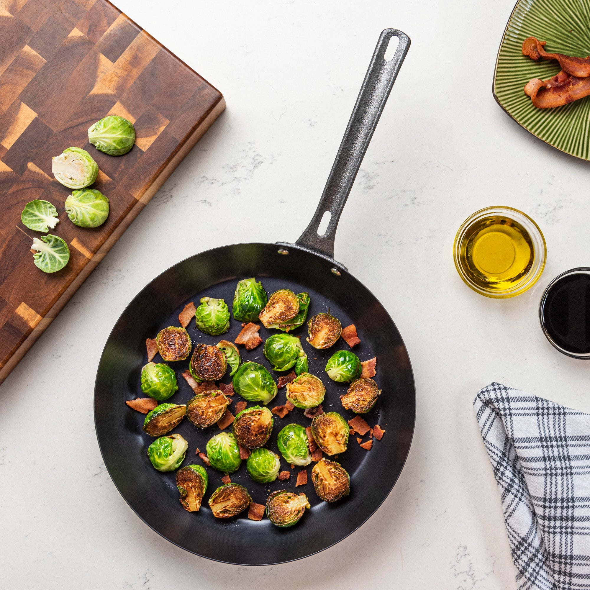 A Viking Culinary Products Blue Carbon Steel 12-Inch Fry Pan with roasted Brussels sprouts and bacon sits on a white countertop, near a cutting board with cut sprouts, a green plate of bacon, oil in a glass, and a checkered towel.