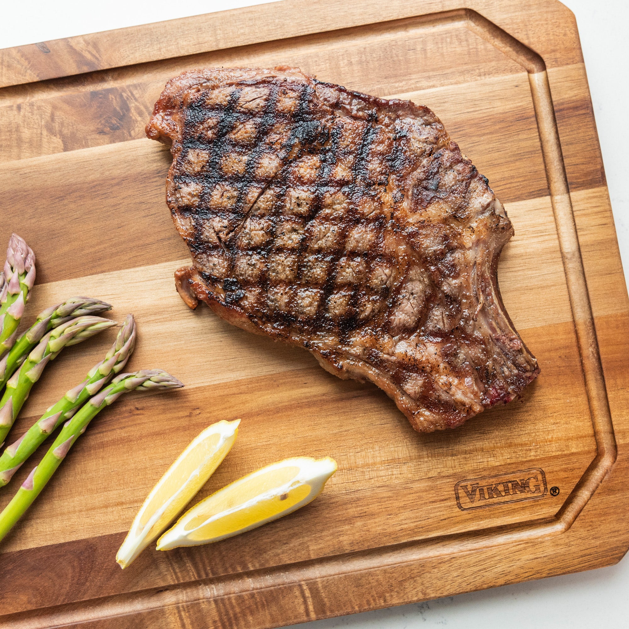 Grilled steak with crosshatch grill marks on the Viking Acacia Carving Board with Juice Groove by Viking Culinary Products, served alongside asparagus spears and two lemon wedges.