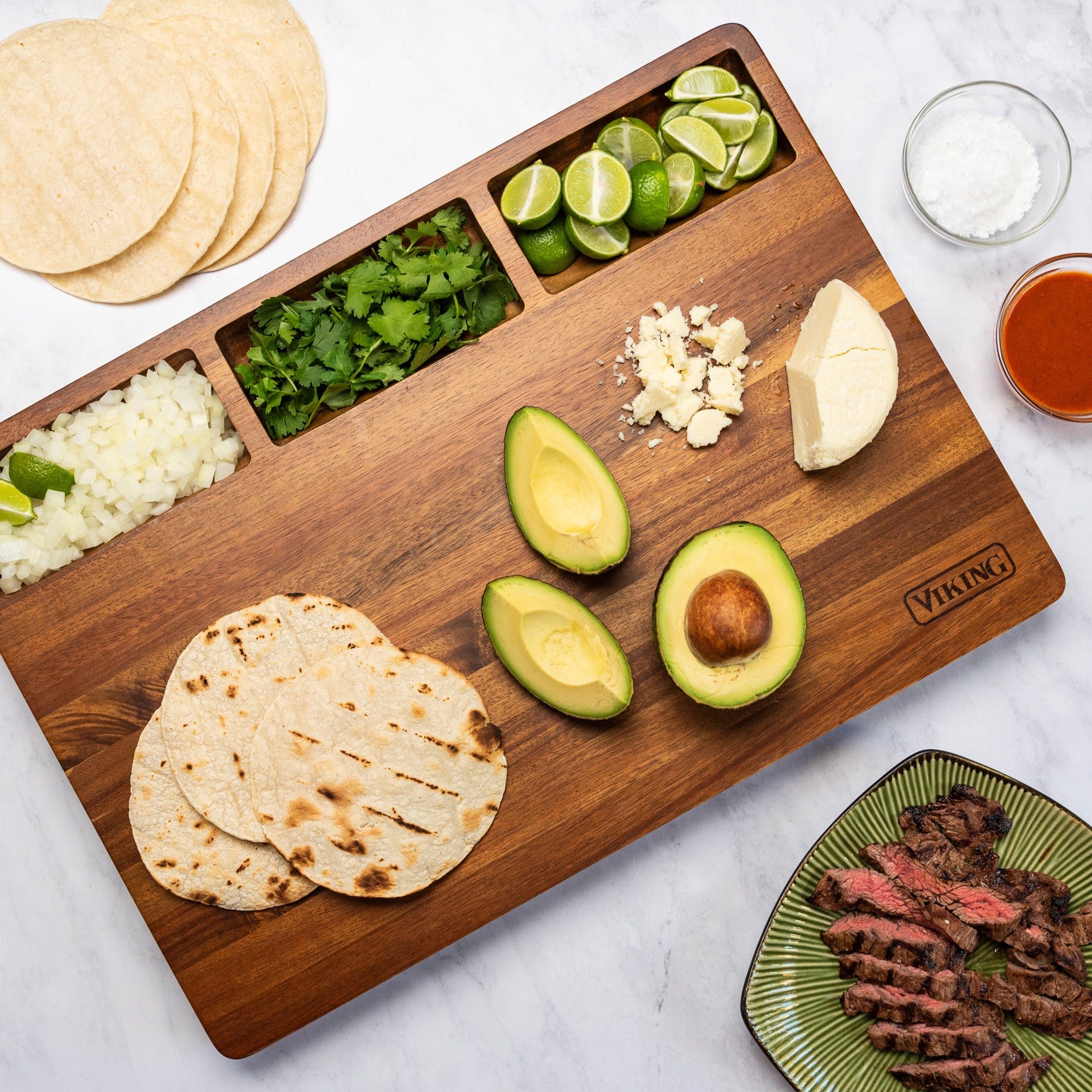 The Viking Acacia 20-Inch Reversible Butcher Block Prep and Carving Board by Viking Culinary Products holds tortillas, onions, cilantro, limes, avocado halves, crumbled cheese, and sauce; grilled steak is served on a green plate with a small salt bowl nearby.