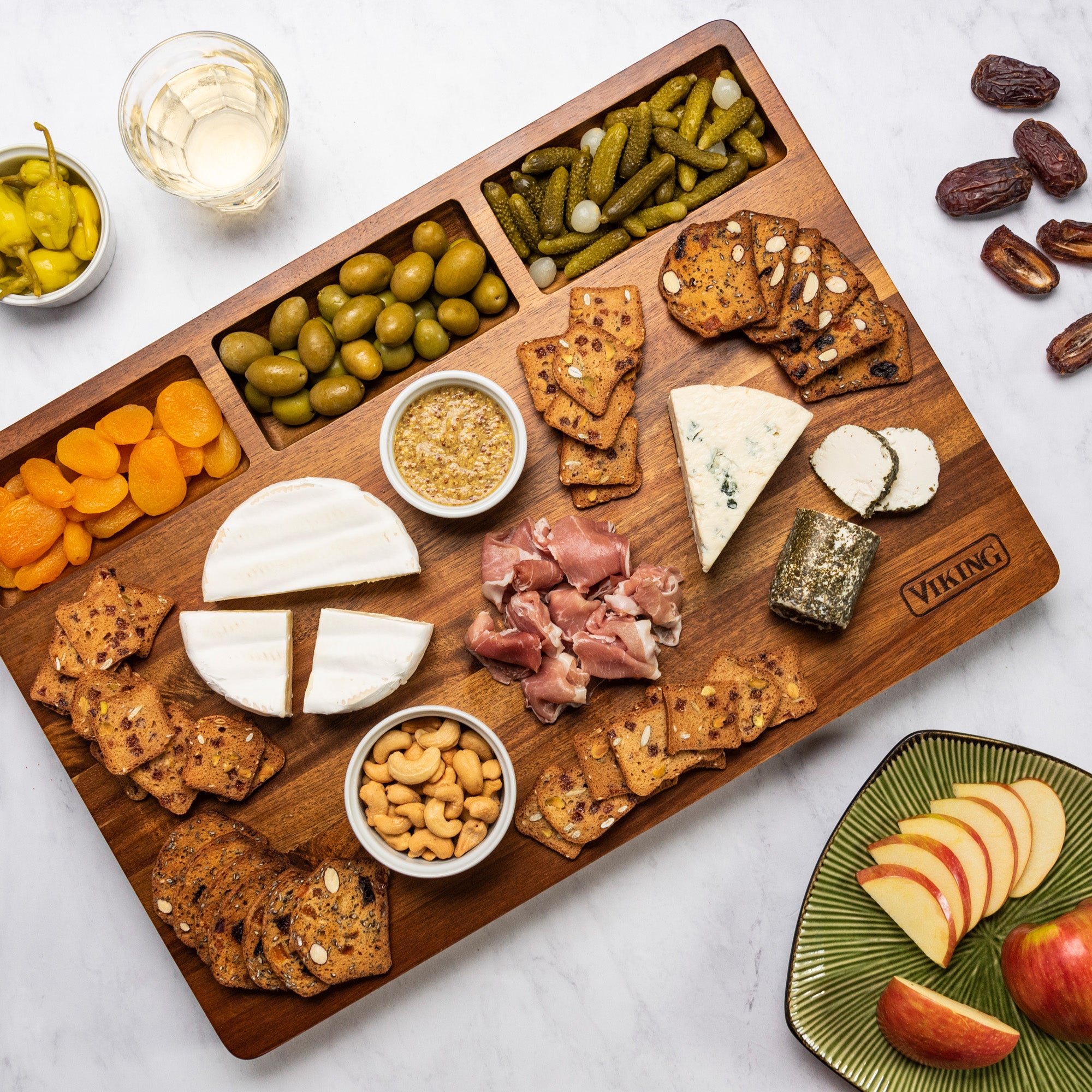 The Viking Acacia 20-Inch Reversible Butcher Block Prep and Carving Board by Viking Culinary Products displays cheese, crackers, olives, dried apricots, pickles, prosciutto, nuts, and mustard beside a green plate of apple slices and a glass of white wine.