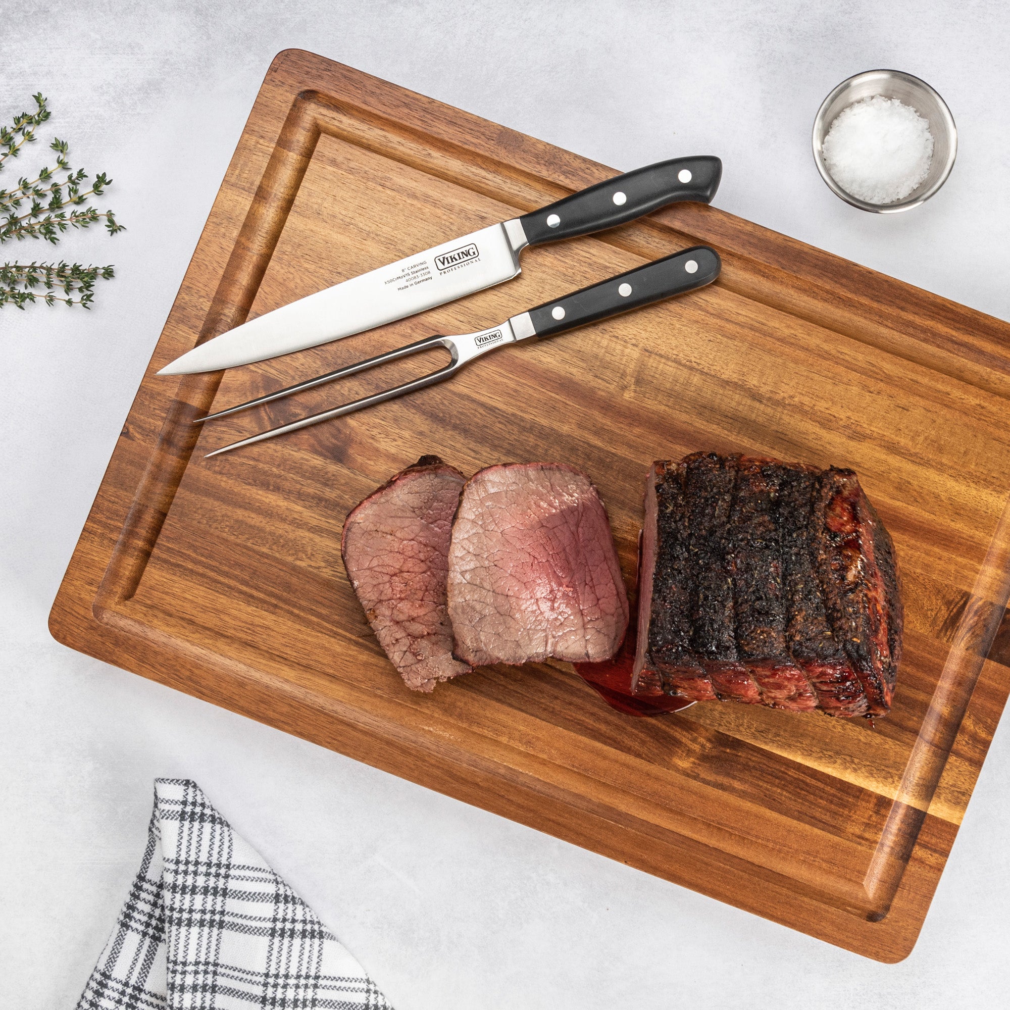 A sliced roast beef rests on a wooden cutting board with the Viking Professional 2-Piece Carving Set by Viking Culinary Products, alongside a small bowl of salt, fresh herbs, and a checkered towel.