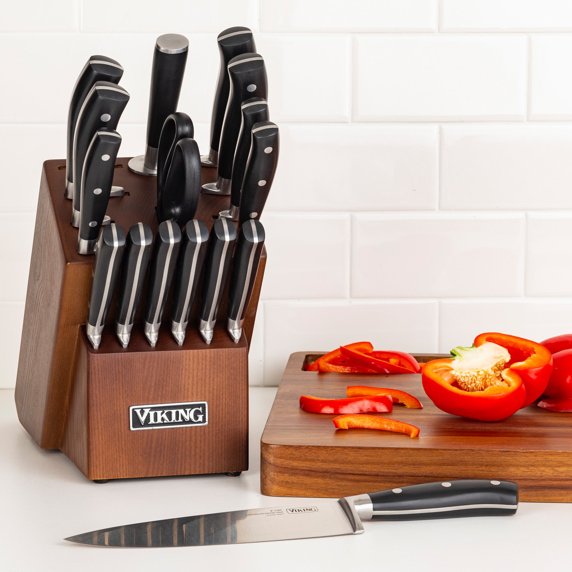 The Viking 17-Piece Cutlery Set with Light Walnut Color Block by Viking Culinary Products, including chef knives of X50CrMoV15 stainless steel and kitchen shears, rests on a counter beside a cutting board with cut and whole red bell peppers against white tiles.