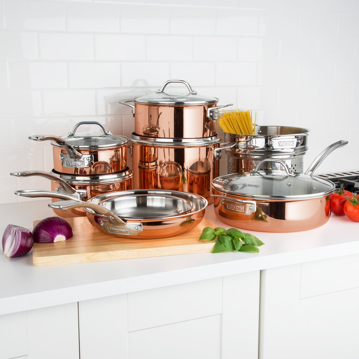 The Viking 3-Ply Copper Clad 13-Piece Cookware Set from Viking Culinary Products, with silver handles and glass lids, is displayed on a kitchen counter near tomatoes, basil, onion, and uncooked spaghetti against a white tile background.