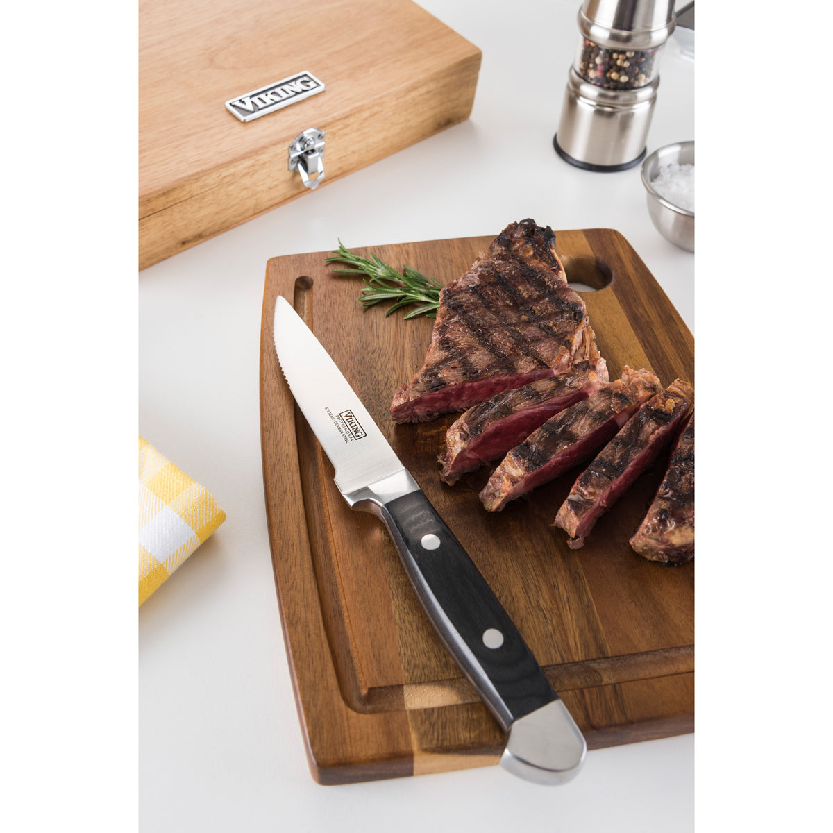 A grilled steak sliced with a Viking Steakhouse Pakka Wood 6-Piece Steak Knife by Viking Culinary Products sits on a wooden board, garnished with rosemary. Nearby are the gift box, pepper grinder, salt bowl, and yellow napkin on a white surface.