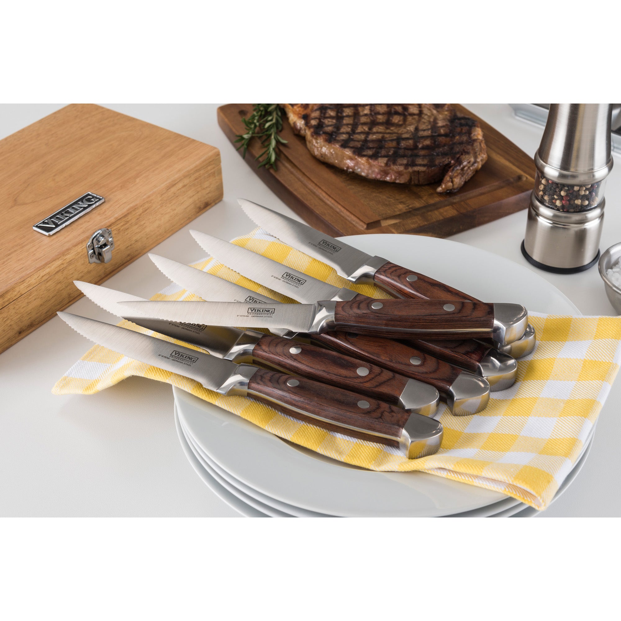 The Viking Steakhouse Pakka Wood 6-Piece Steak Knife Set with Gift Box (Red) by Viking Culinary Products features corrosion-resistant knives neatly arranged on plates with a yellow checkered napkin, steak, a pepper grinder, and a wooden knife box.