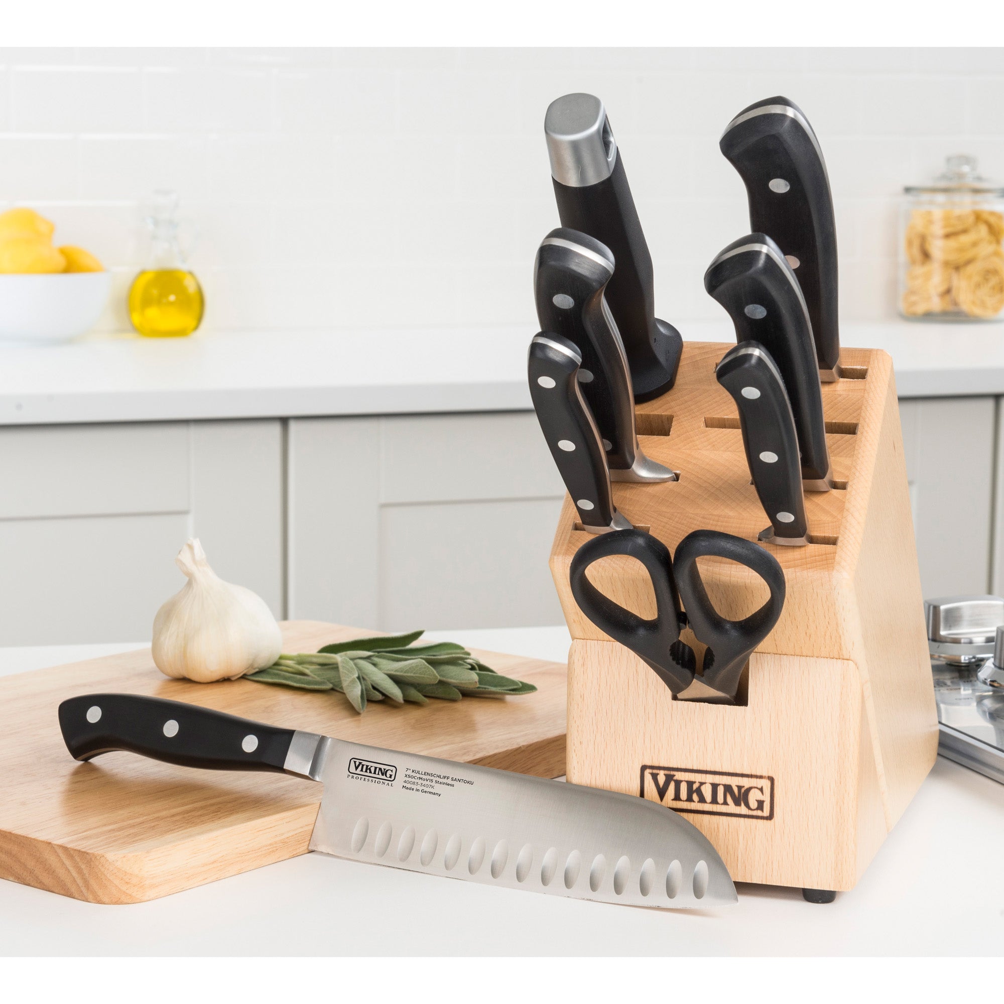 The Viking Professional 7-Piece Cutlery Set by Viking Culinary Products, featuring German steel knives and scissors in a wooden block, is showcased on a cutting board with a chef knife, garlic, and herbs in a modern white kitchen.