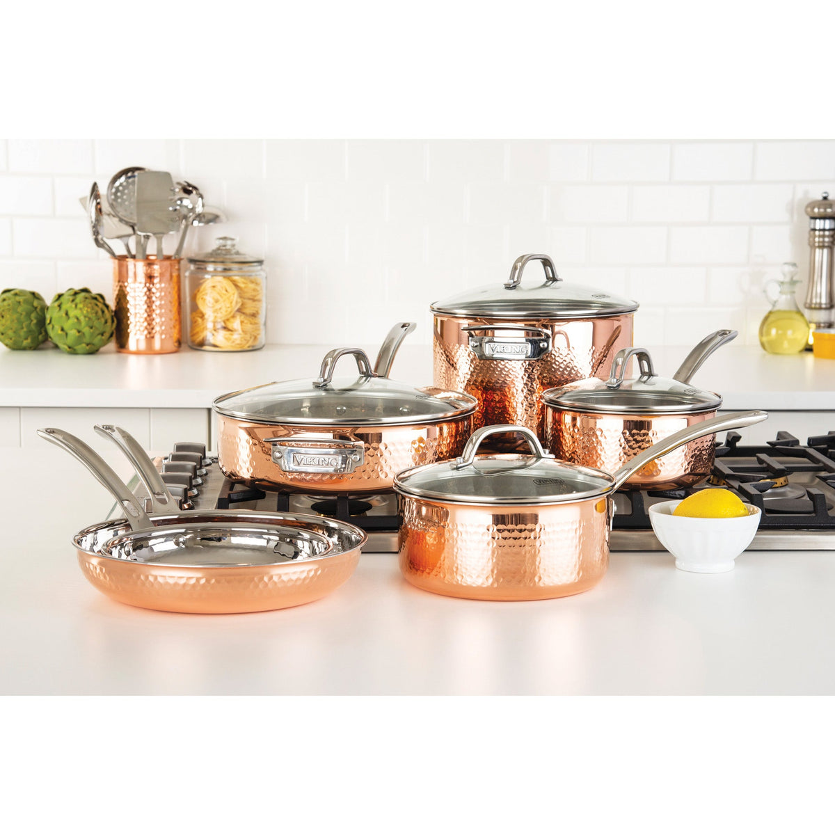 The Viking 3-Ply Hammered Copper Clad 10-Piece Cookware Set with Glass Lids by Viking Culinary Products is displayed on a white kitchen counter and stove, surrounded by utensils, artichokes, pasta jars, and a lemon.