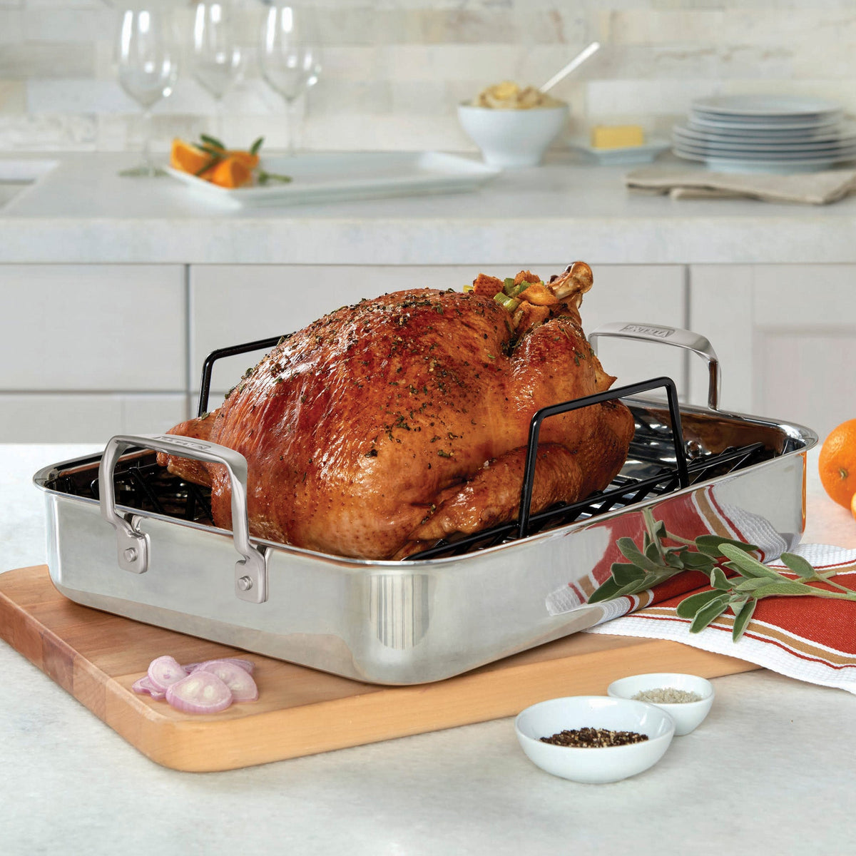 A roasted turkey sits in a stainless steel roasting pan on a kitchen counter, with herbs on top. Plates, glasses, spices, and a bowl of butter are visible in the background.