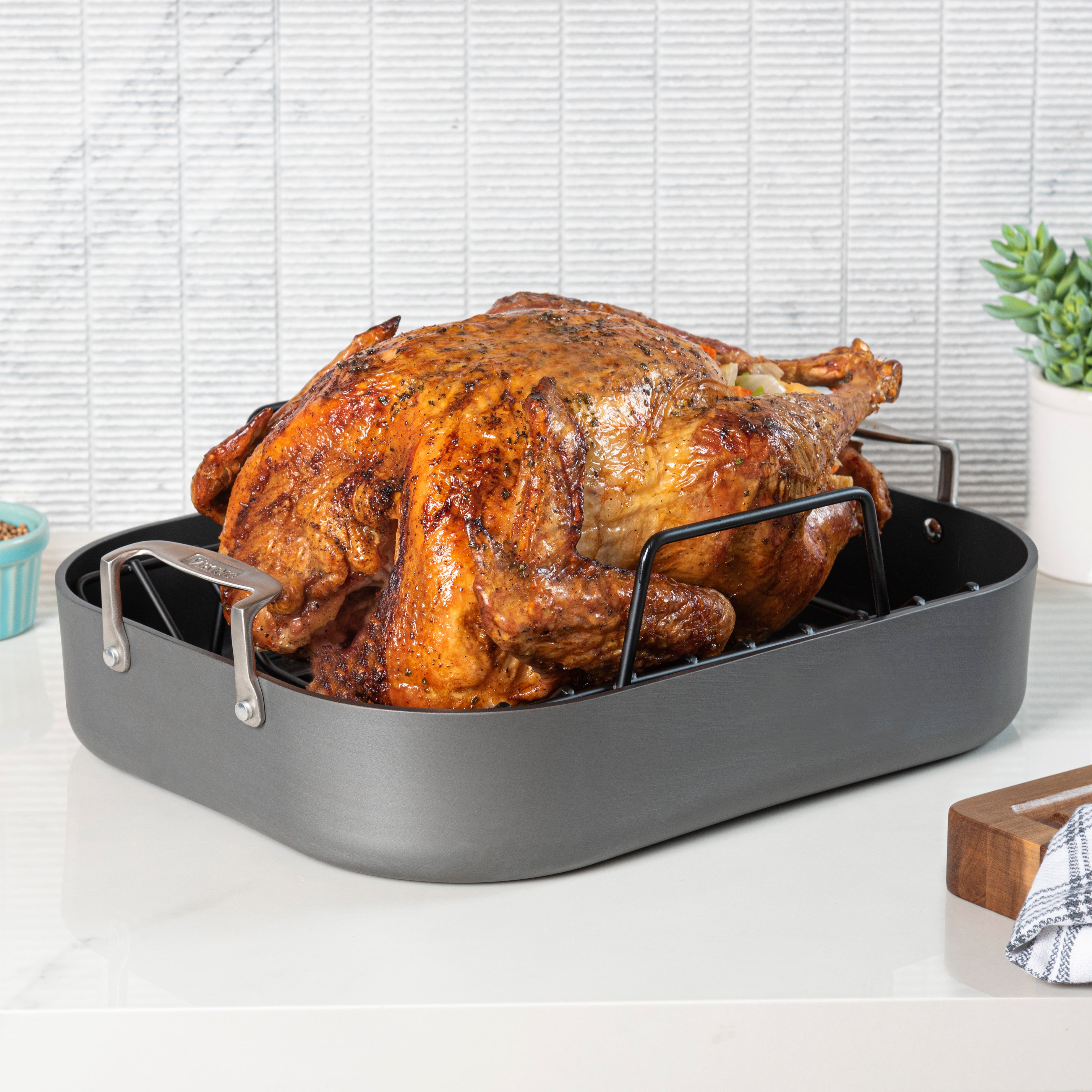 A golden-brown roasted turkey sits in a large gray roasting pan with a rack on a white countertop, with a plant and kitchen items in the background.