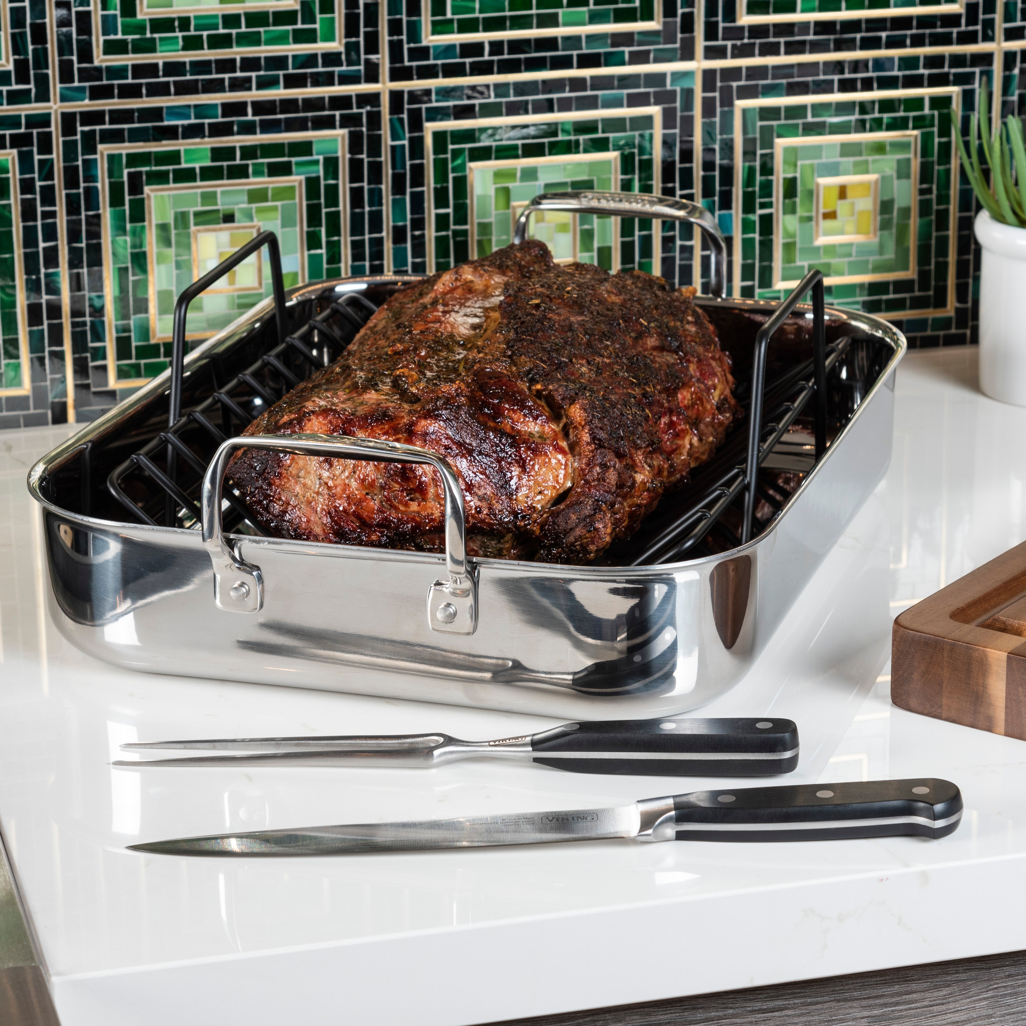 A roasted beef brisket sits in a stainless steel roasting pan on a kitchen counter, with a carving fork and knife placed in front, and a green tiled backsplash in the background.