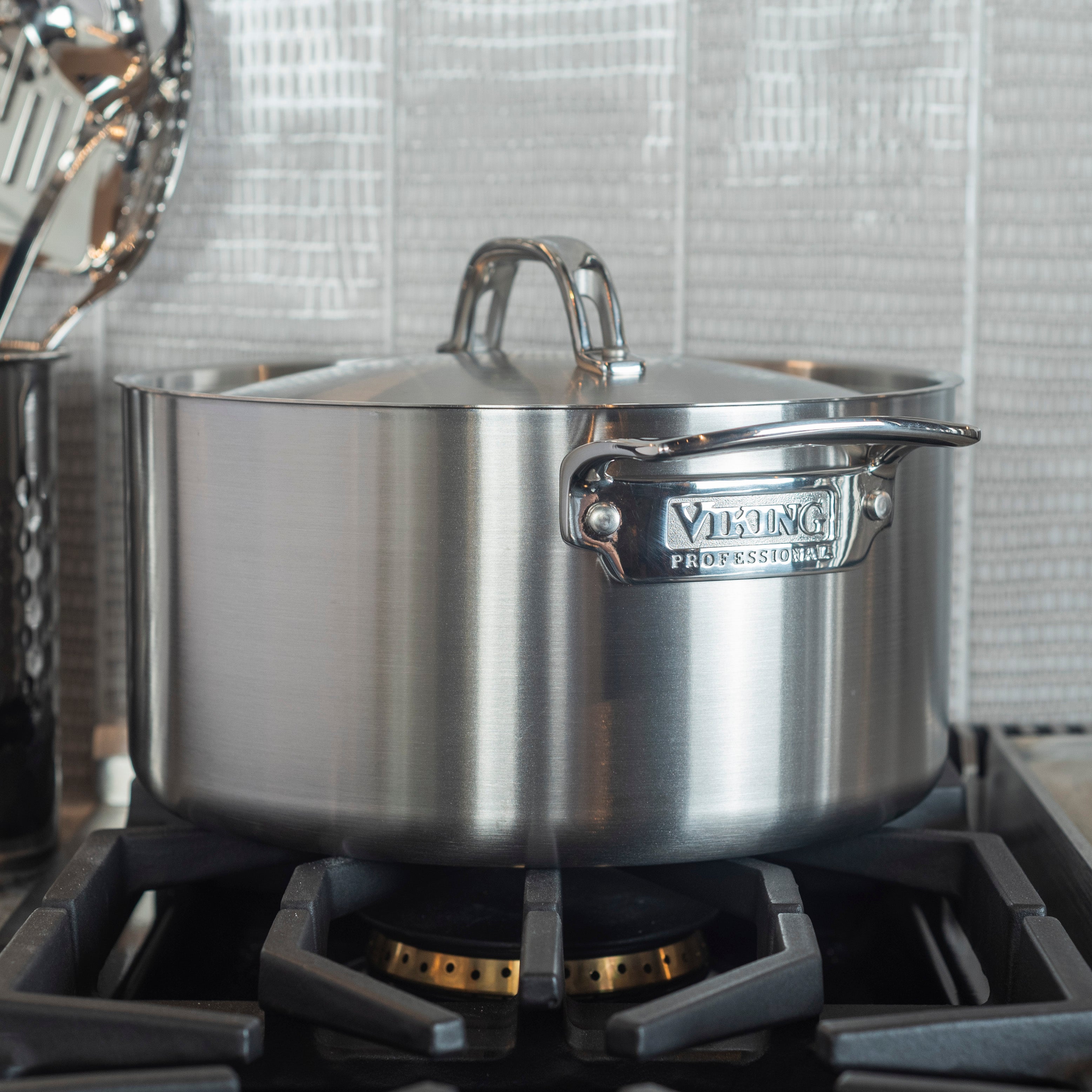 A stainless steel Viking Professional pot with a lid sits on a gas stove burner in a modern kitchen, with utensils and a textured tile backsplash visible in the background.