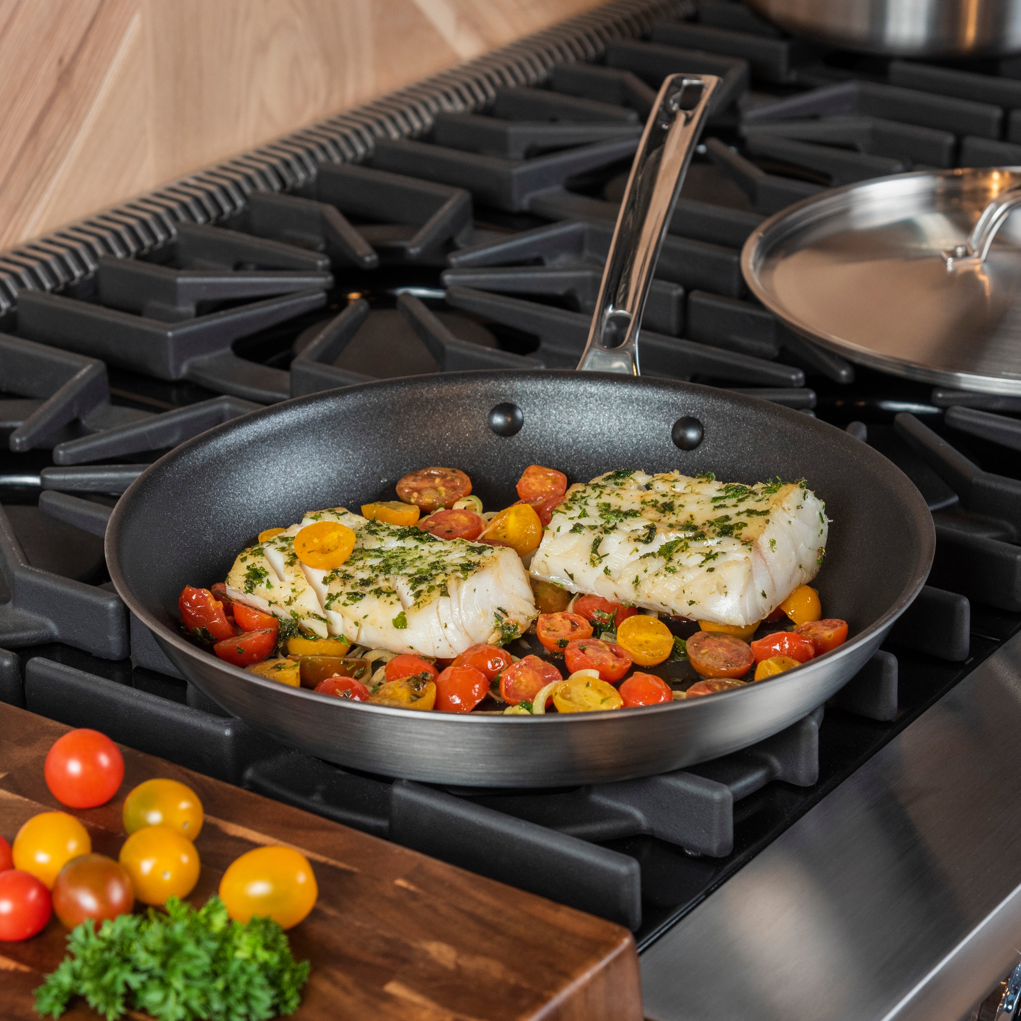 Two pieces of fish topped with herbs are cooking in a skillet with halved red and yellow cherry tomatoes on a stovetop; a cutting board with more tomatoes is nearby.