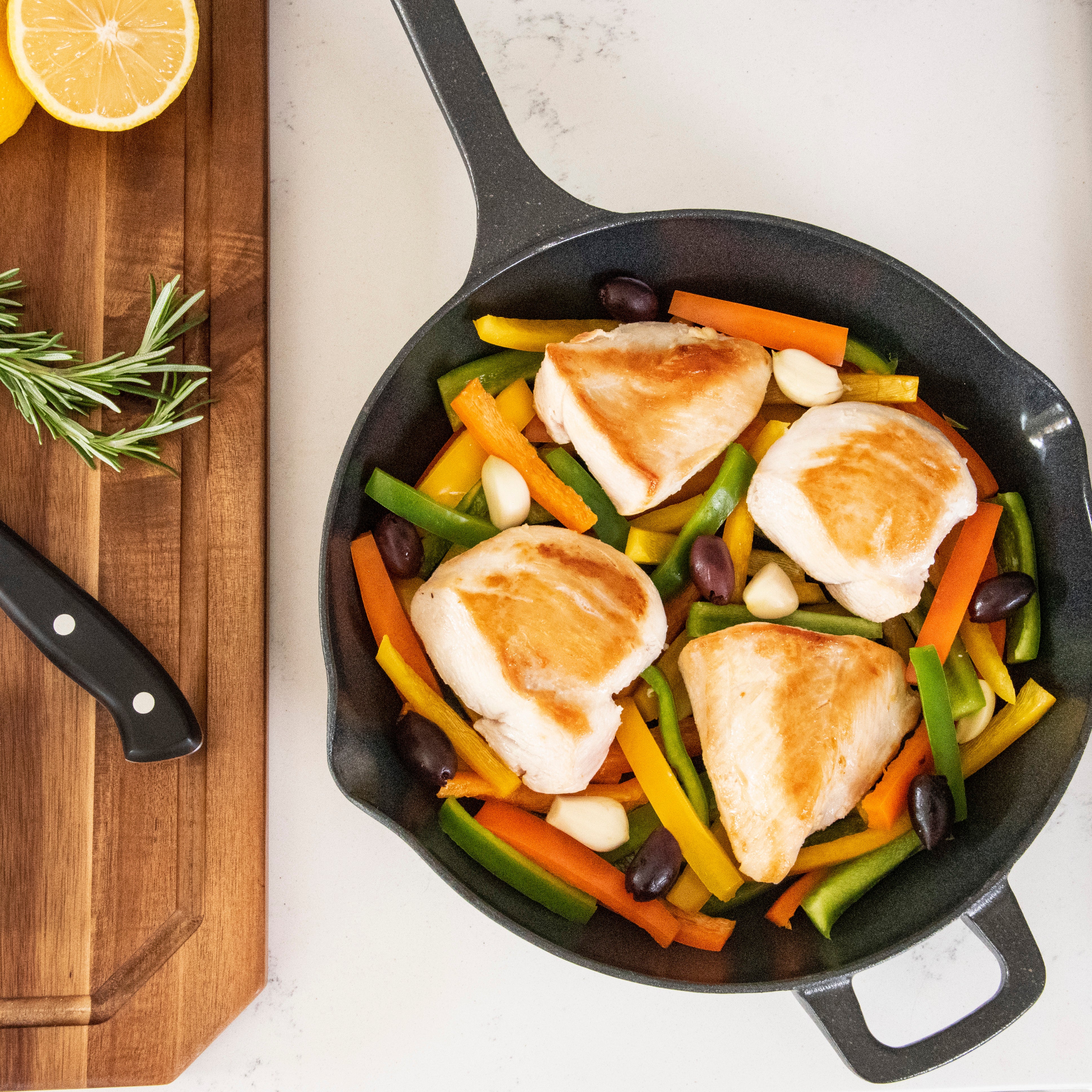 A skillet with browned chicken breasts, sliced bell peppers, garlic cloves, and olives. Nearby, a cutting board holds a halved lemon, fresh rosemary, and a knife.