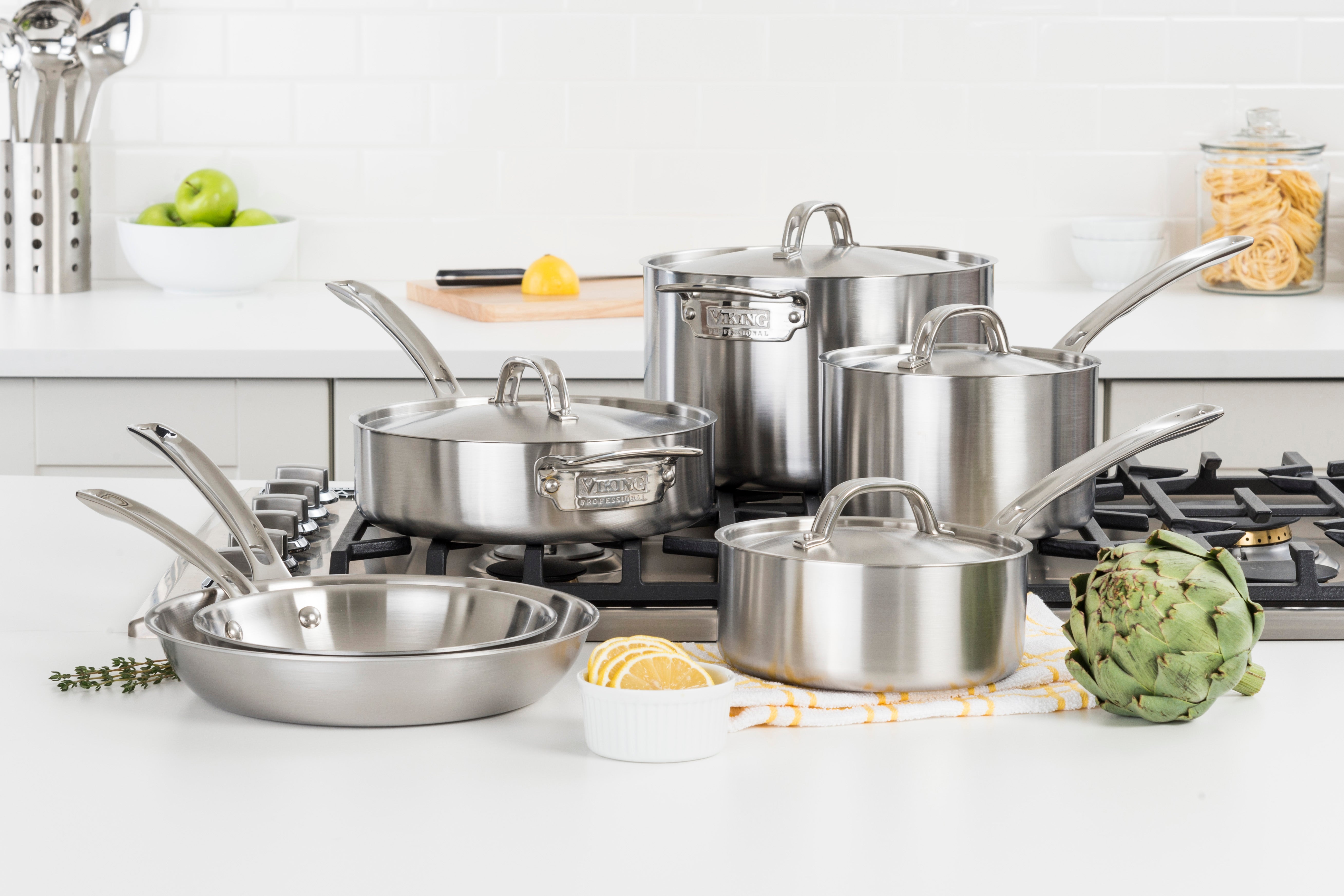 A set of stainless steel pots and pans with lids arranged on a kitchen stove and counter, with an artichoke, sliced lemon, herbs, and kitchen utensils in the background.