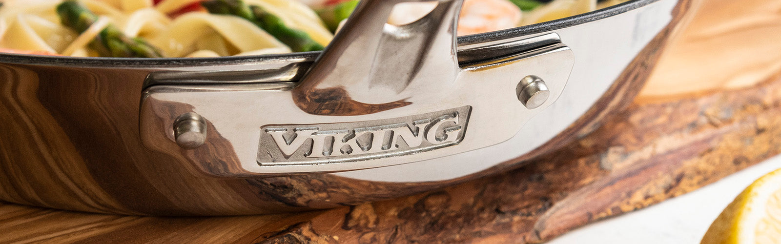Close-up of a shiny Viking stainless steel pot handle with pasta and vegetables inside. The pot rests on a wooden cutting board, and a lemon wedge is partially visible in the corner.