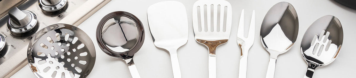 A row of shiny stainless steel kitchen utensils, including a slotted spoon, ladle, spatula, fork, and serving spoons, are arranged neatly on a countertop.