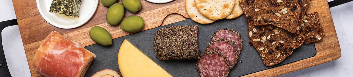 A charcuterie board with assorted cheeses, cured meats, green olives, crackers, and seeded crisps, arranged on a wooden board with a slate serving section.