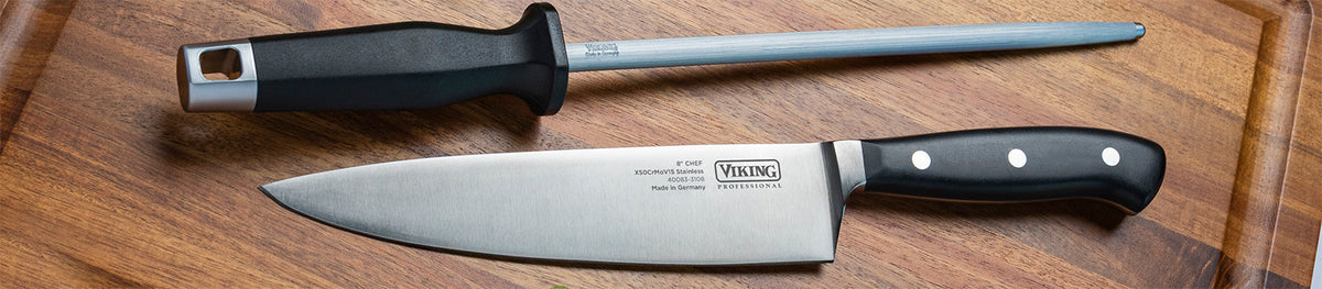 A chef’s knife and a honing steel with black handles rest on a wooden cutting board.