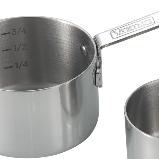 A stainless steel saucepan with measurement markings for 1/4, 1/2, and 3/4 on the inside and a handle labeled VIKING. Another similar pot is partially visible next to it.