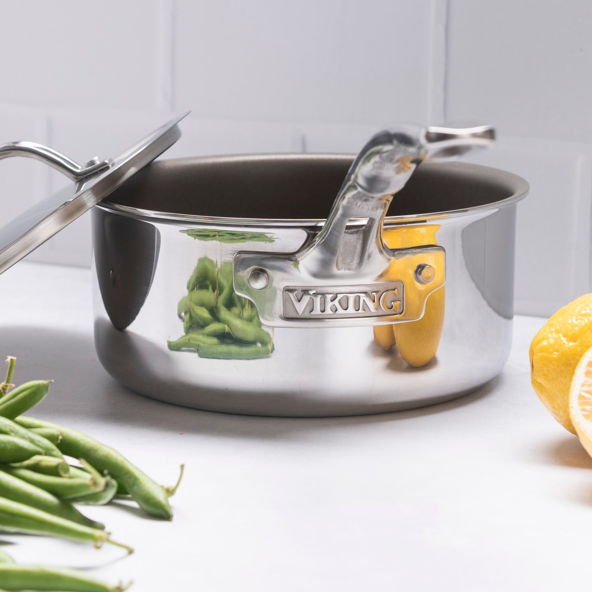 The Viking PureGlide Pro 3-Ply 3-Quart Sauce Pan with Metal Lid by Viking Culinary Products, featuring a shiny stainless steel finish and chemical-free nonstick interior, sits on a white surface near green beans and a sliced lemon.