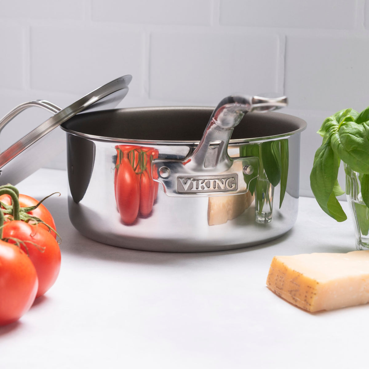 The Viking Culinary Products PureGlide Pro 3-Ply 2-Quart Sauce Pan with Metal Lid sits on a white surface, surrounded by fresh tomatoes, cheese, and basil, with a white tile background—perfect for healthy cooking.
