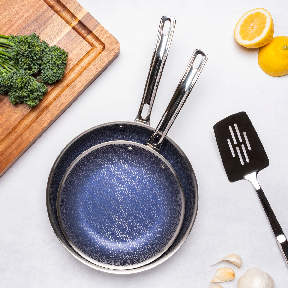 The Viking 3-Ply Hybrid Plus Ceramic 2 Pc Nonstick Fry Pan Set by Viking Culinary Products, featuring 9.5-inch and 11-inch pans, nests neatly beside a cutting board with broccolini, spatula, garlic, and lemons—ideal for induction kitchens.