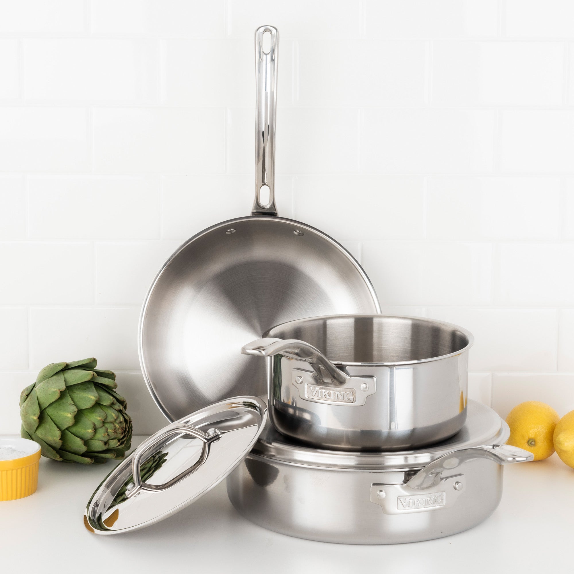 The Viking 3-Ply Stainless Steel 5-Piece Cookware Set with Metal Lids by Viking Culinary Products is stacked on a white countertop, with an artichoke, a yellow ramekin, and two lemons nearby against a white tile backsplash.