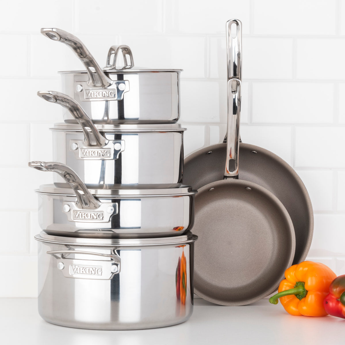 A Viking Culinary Products PureGlide Pro 3-Ply 10-Piece Cookware Set and two chemical-free nonstick frying pans sit stacked on a white kitchen surface with yellow and red bell peppers, backed by a white tiled backsplash.