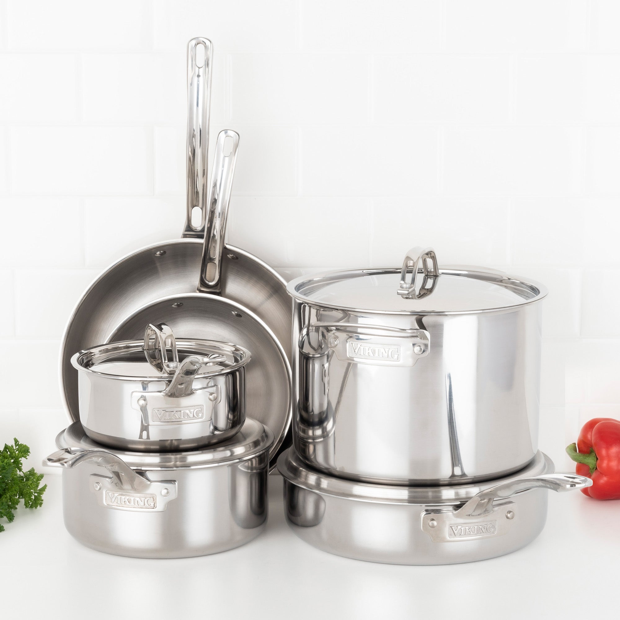 The Viking Culinary Products 3-Ply Stainless Steel 10-Piece Cookware Set with metal lids, suitable for all cooking surfaces, is displayed on a white kitchen countertop with parsley and a red bell pepper adding color.