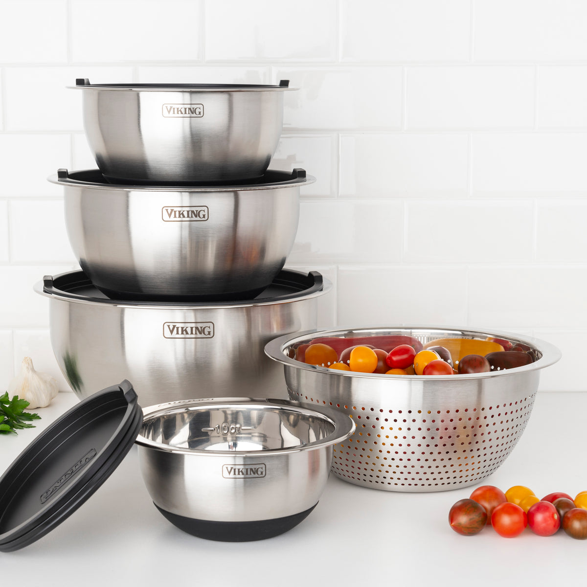 The Viking 9-Piece Stainless Steel Mixing Bowl Set with Strainer by Viking Culinary Products features four stacked bowls with airtight lids, a metal strainer holding cherry tomatoes, and a black silicone base on a white kitchen counter.