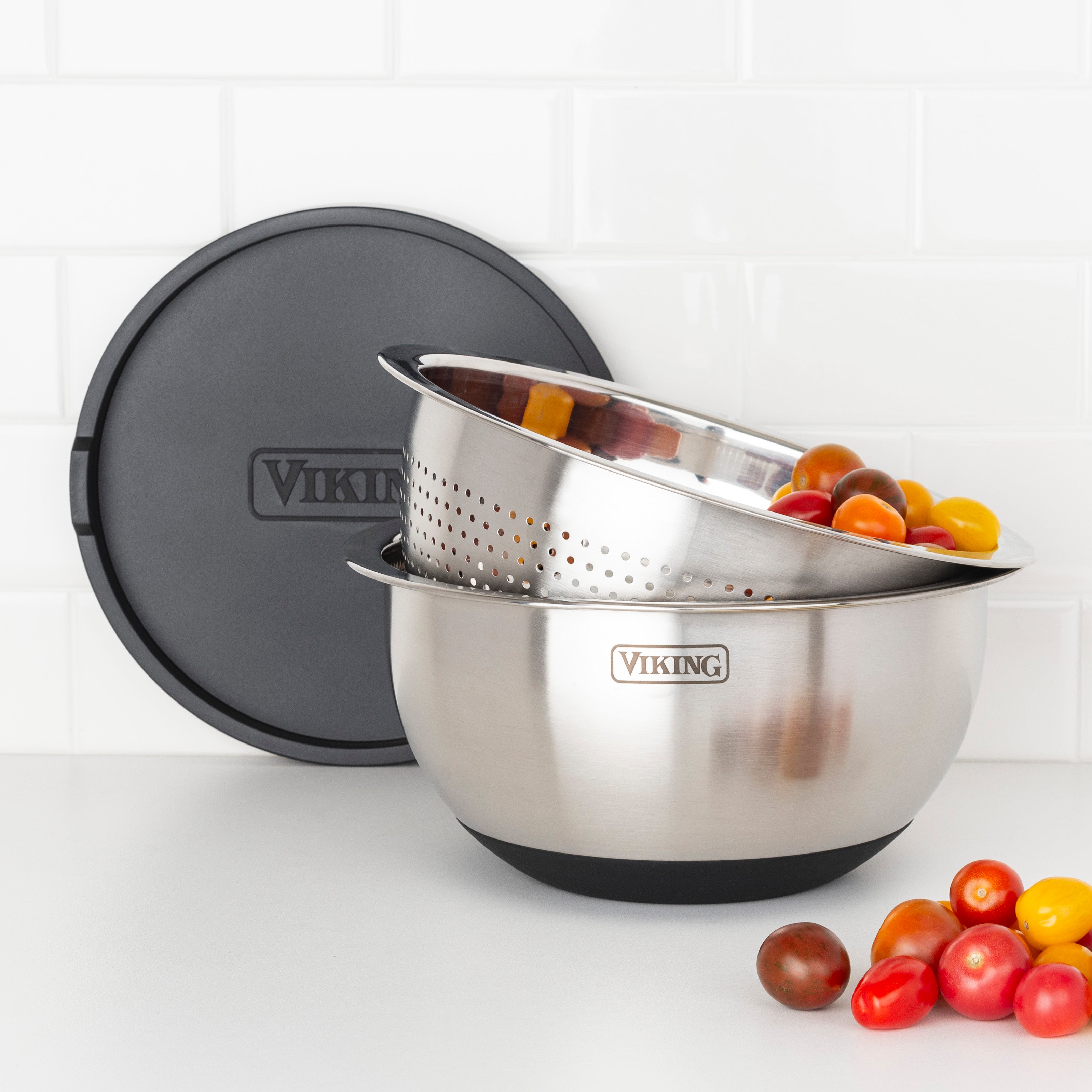 A Viking Culinary Products 9-Piece Stainless Steel Mixing Bowl Set with Strainer and black airtight lid features a colander of colorful tomatoes, with more scattered on a white countertop. The bowl has a silicone base for stability.