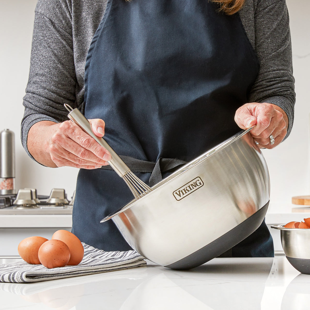 A person wearing a dark apron whisks ingredients in a Viking Culinary Products 9-Piece Stainless Steel Mixing Bowl with Strainer and black silicone base, on a kitchen counter with eggs and a striped towel nearby.