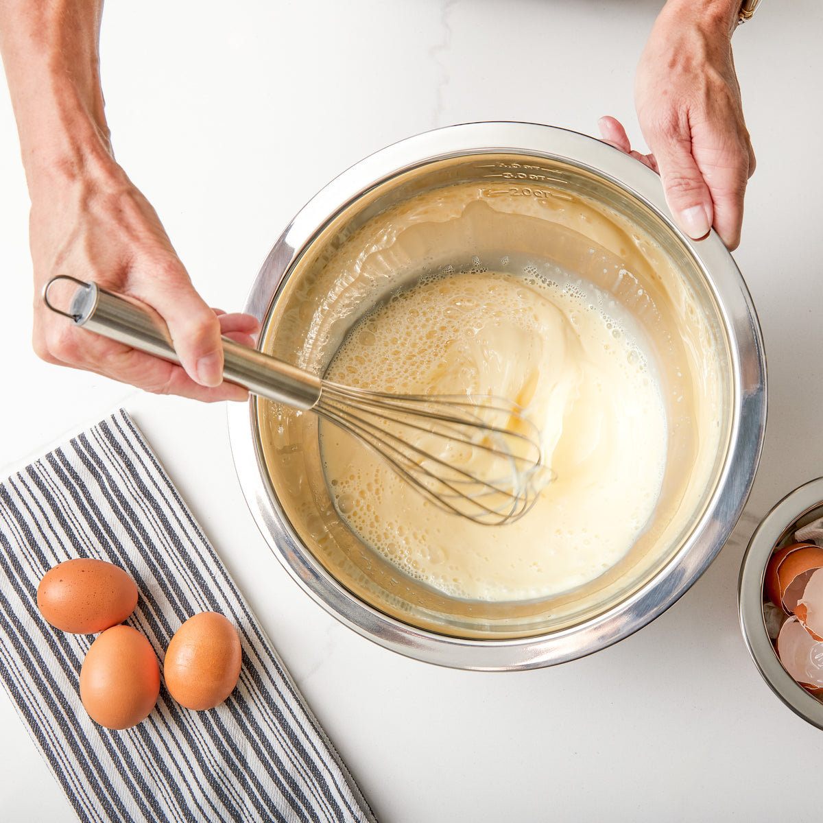 A person whisks a creamy mixture in a Viking Culinary Products Viking 9-Piece Stainless Steel Mixing Bowl with Strainer, Black, beside a striped towel, three brown eggs, and eggshells on a white countertop.