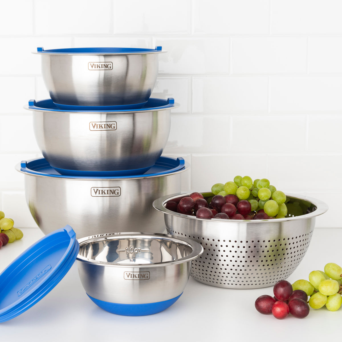 The Viking 9-Piece Stainless Steel Mixing Bowl Set with Strainer, Blue by Viking Culinary Products is stacked on a countertop beside a colander of grapes, with blue airtight lids and silicone bases. Grapes are scattered around the bowls.