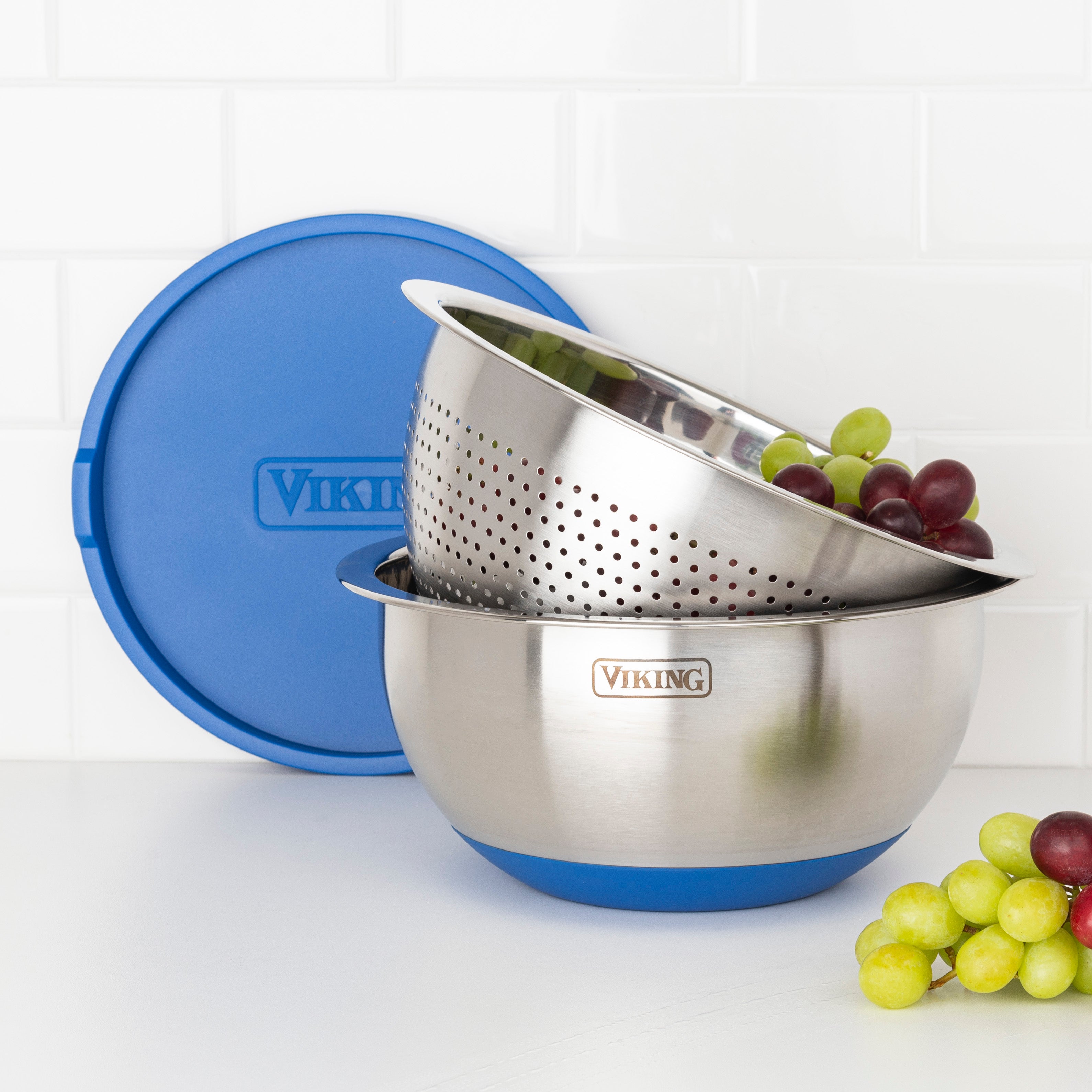 The Viking 9-Piece Stainless Steel Mixing Bowl Set with Strainer by Viking Culinary Products features a colander holding grapes inside a matching bowl, both with blue silicone bases, and an airtight lid resting on a white kitchen countertop.
