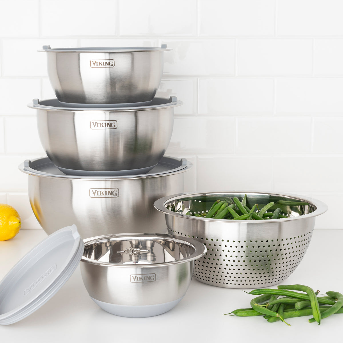 The Viking 9-Piece Stainless Steel Mixing Bowl Set with Strainer by Viking Culinary Products is neatly stacked on a white counter, beside a metal strainer filled with green beans. Loose green beans and a lemon add to this compact kitchen storage display.