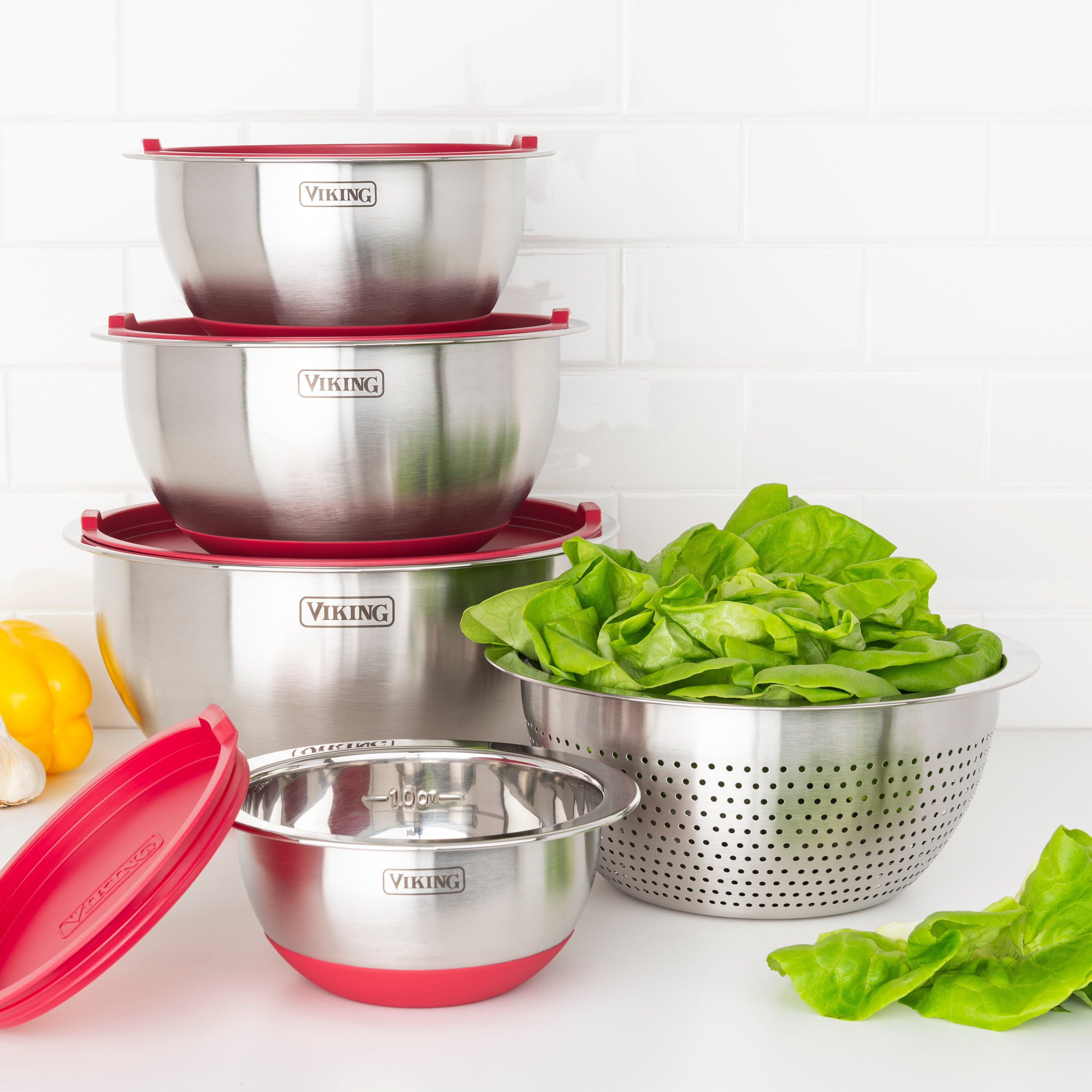 The Viking Culinary Products 9-Piece Stainless Steel Mixing Bowl Set with Strainer in red is stacked compactly on the kitchen counter beside a colander of fresh spinach, with a white subway tile backsplash in the background.