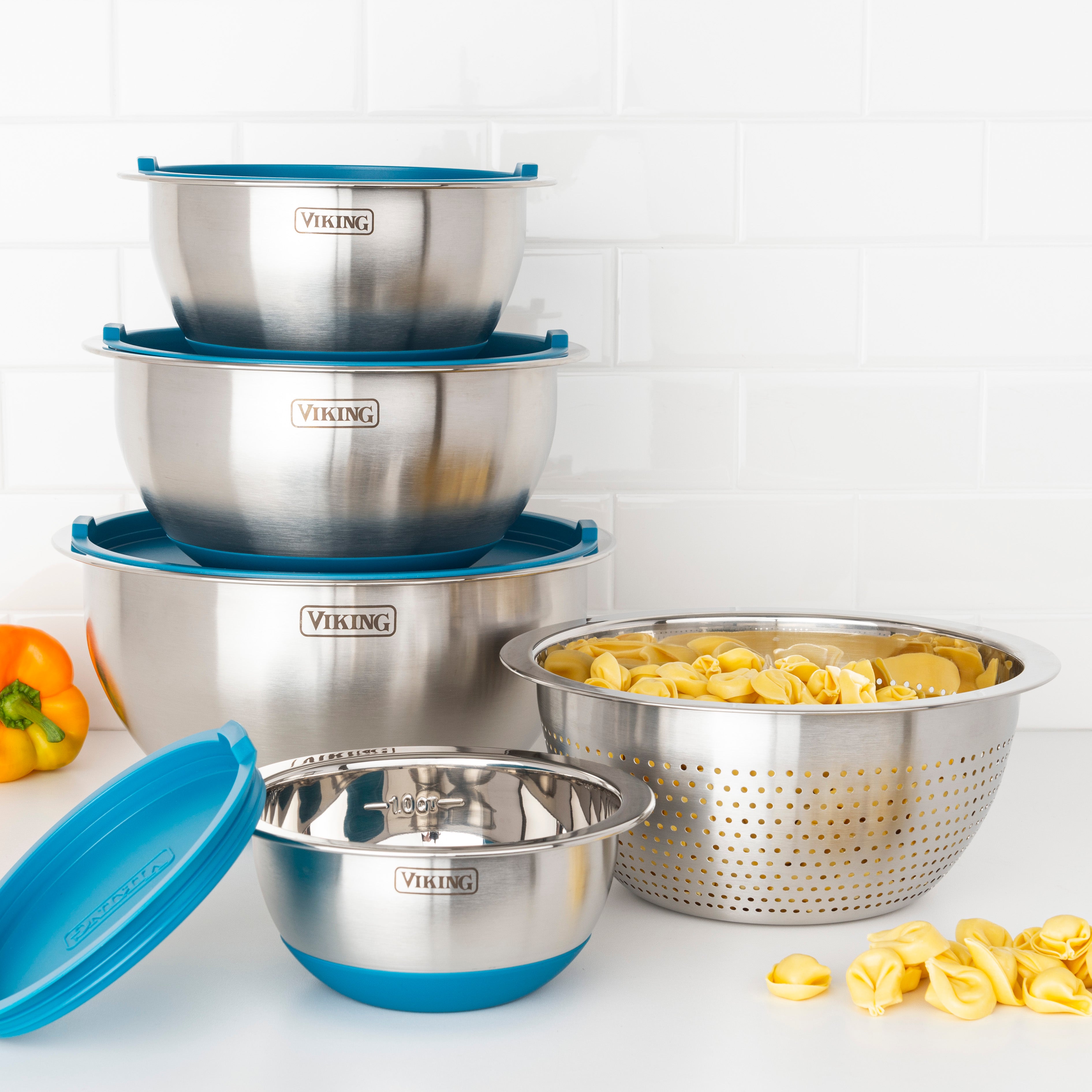 The Viking 9-Piece Stainless Steel Mixing Bowl Set with Strainer by Viking Culinary Products is stacked on a white counter beside a colander of tortellini and a yellow bell pepper, all set against a white tile backsplash—ideal for compact storage.