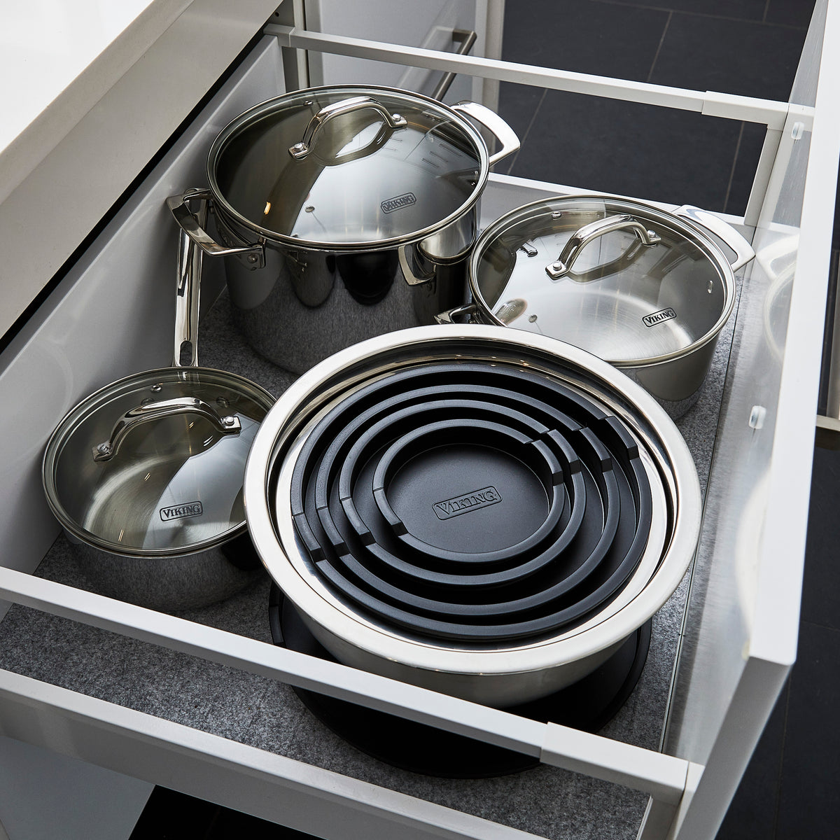 Open kitchen drawer containing three stainless steel pots with lids and a set of nested black and stainless steel pans. The pots are organized neatly on a gray liner inside the white drawer.