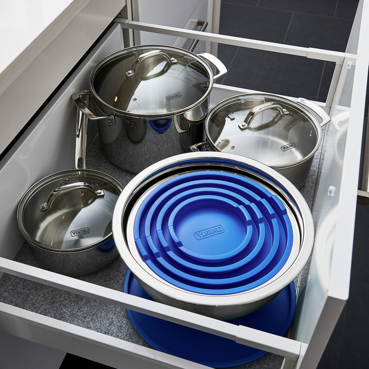 Open kitchen drawer with neatly organized stainless steel pots and pans. Several blue collapsible mixing bowls are stacked inside a large metal bowl on the right side. The drawer has a clean, modern appearance.