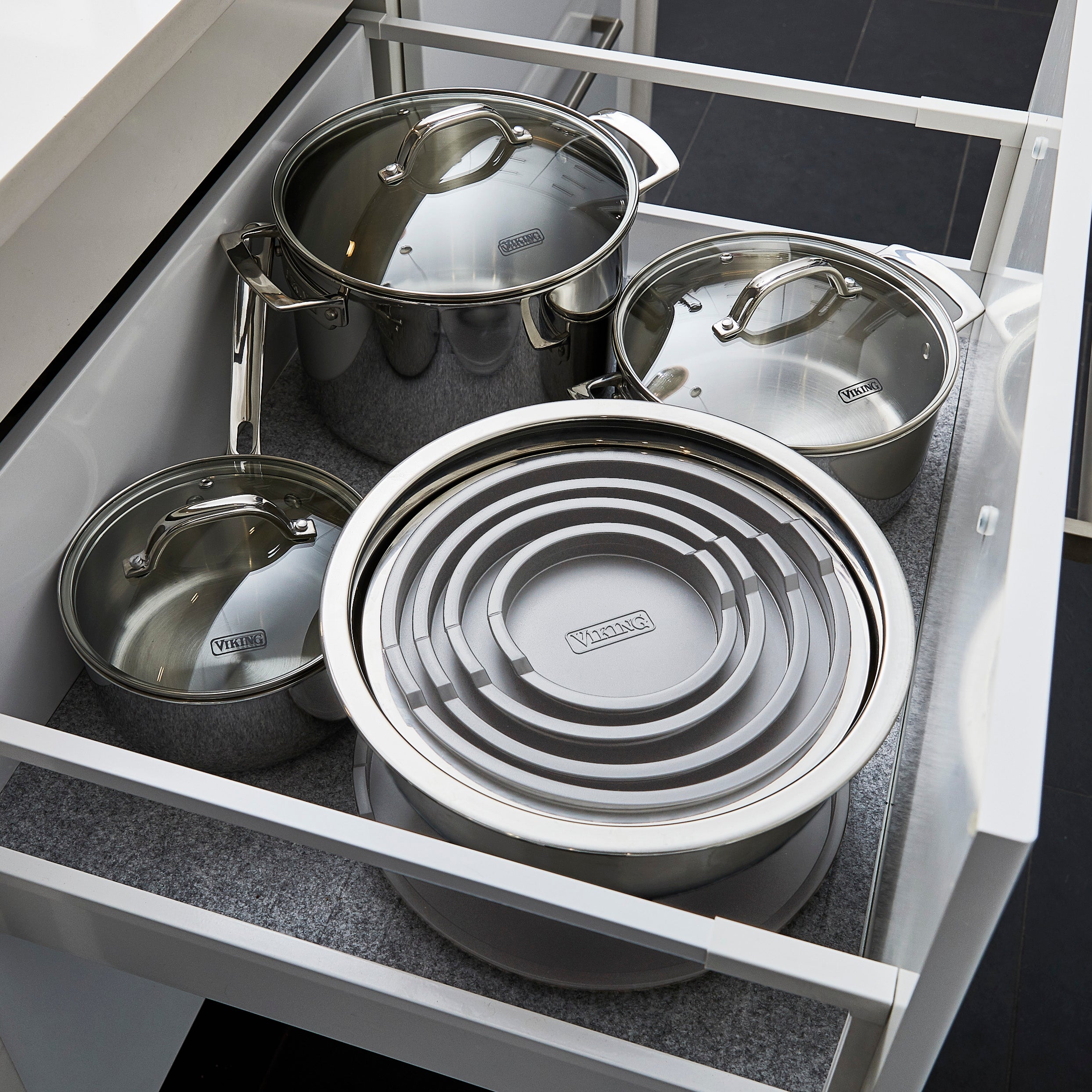 Open kitchen drawer containing three stainless steel pots with lids and a set of nested, circular, gray Viking brand mixing bowls with lids. The drawer has a gray liner, and the kitchen flooring is dark.