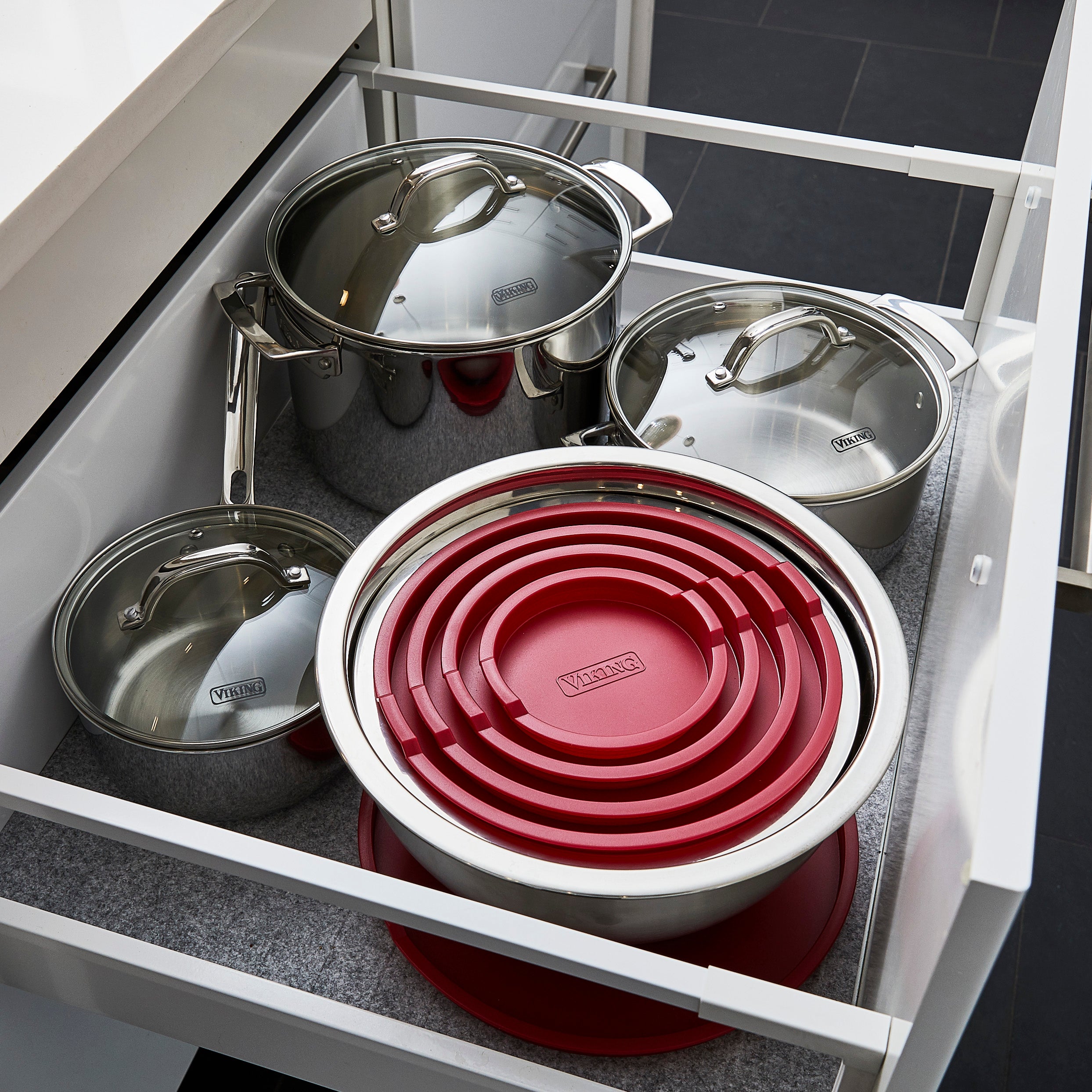 An open kitchen drawer containing stainless steel pots with lids and a set of stainless steel mixing bowls with red lids, neatly nested together. The drawer is lined with a grey mat.