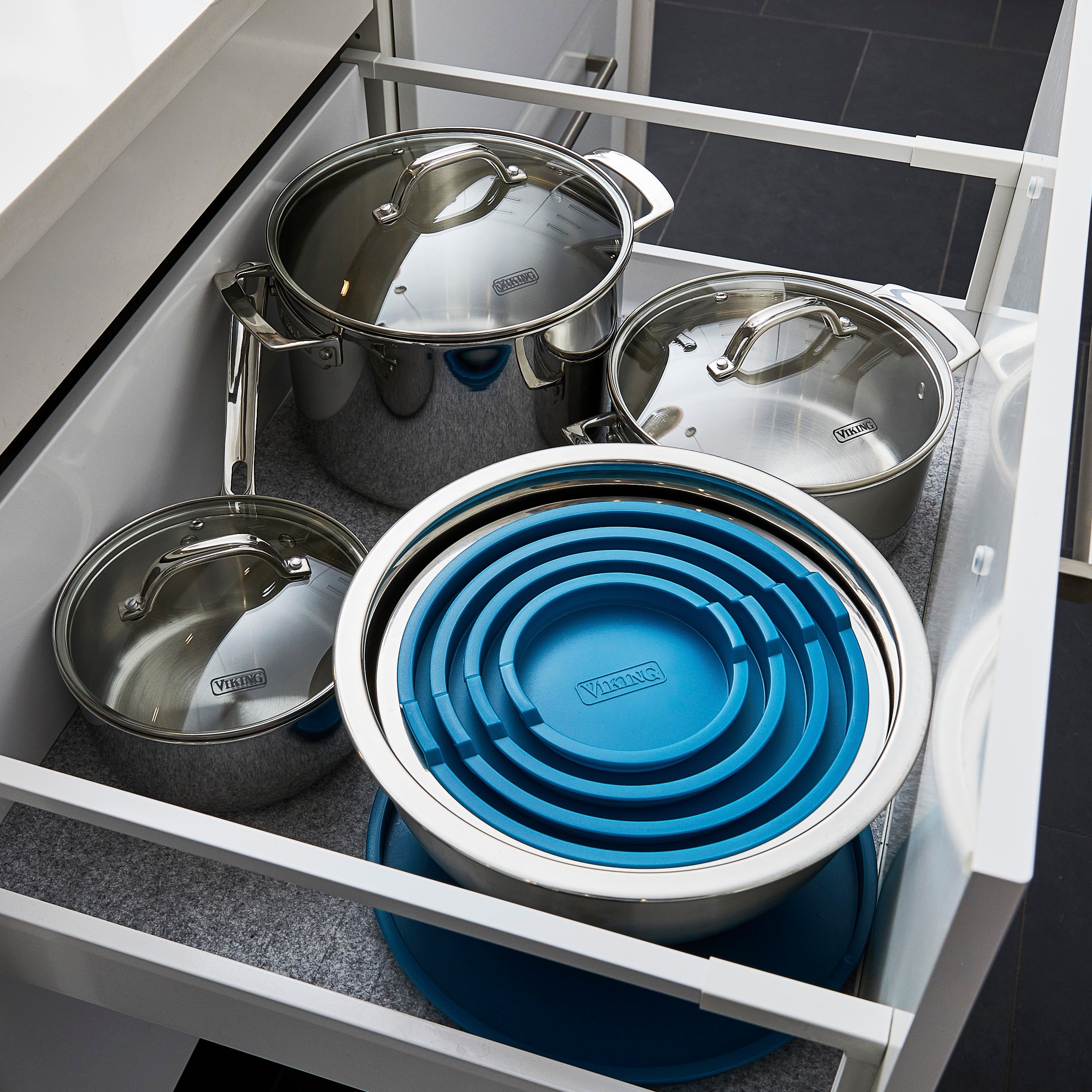 Open kitchen drawer containing three stainless steel pots with lids and a set of stainless steel mixing bowls stacked together with blue silicone lids in various sizes. The drawer has a gray lining on the bottom.