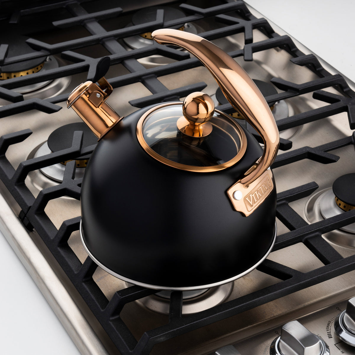 A black and copper tea kettle sits on a modern gas stovetop, featuring a curved handle and spout lever, with burner grates visible around it.