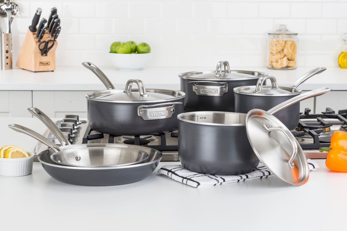 A set of stainless steel pots and pans with lids is displayed on a kitchen counter and stove, surrounded by kitchen items like knives, lemons, a bell pepper, and jars in a modern white kitchen.