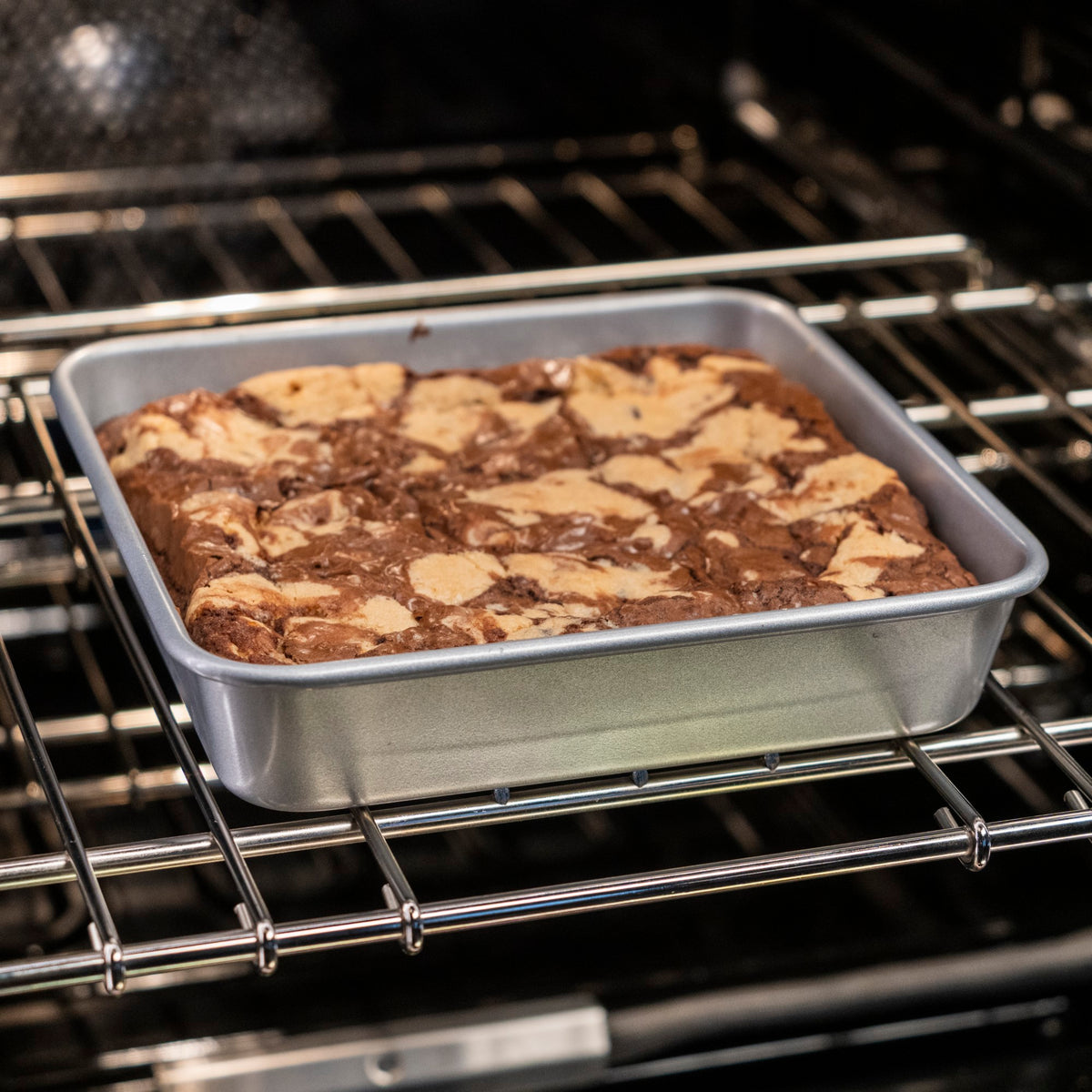 A Viking Culinary Products Aluminized Steel Nonstick 9-inch Square Cake Pan bakes marbled brownies with a chocolate and light batter swirl on the middle oven rack, resting on a commercial-grade bakeware sheet.