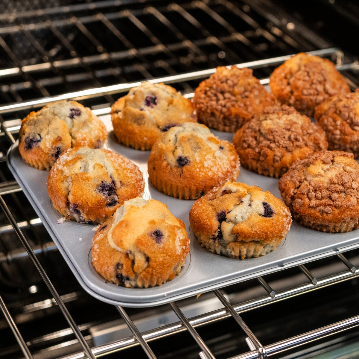 A muffin tin with freshly baked blueberry and streusel-topped muffins sits on an oven rack. The muffins are golden brown and look fluffy and moist.